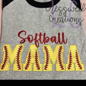 Op de afbeelding: Een grijs en zwart baseballshirt met de woorden "Softball Mama" geborduurd in rood en geel. De letters zijn geel omrand en hebben rode stiksels die lijken op baseballnaden.