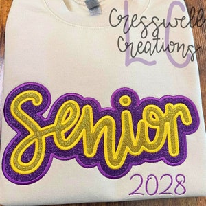 May include: A white sweatshirt with a purple and gold glitter design that says "Senior 2028".