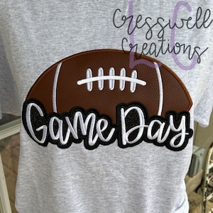 Puede incluir: Una camiseta gris con un bal&oacute;n de f&uacute;tbol marr&oacute;n bordado y el texto "Game Day" en negro y blanco.