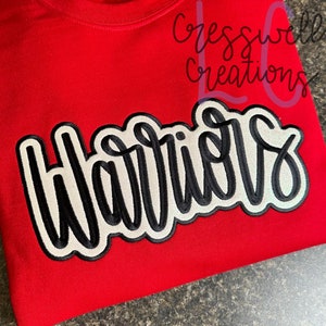 May include: Red sweatshirt with a white and black embroidered patch that says "Warriors". The text is in a cursive font with a black outline.