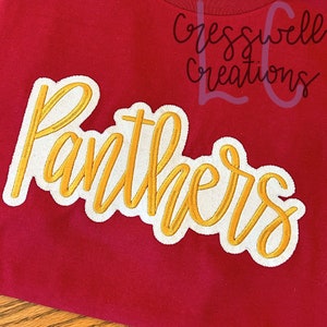 May include: A red t-shirt with the word "Panthers" embroidered in gold script with a white border. The shirt also features the text "Cresswell Creations" in a cursive font.