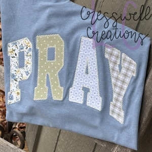 Könnte beinhalten: Hellblaues T-Shirt mit dem Wort "PRAY", das in floralen, gepunkteten und karierten Mustern appliziert ist. Das Shirt zeigt auch den Text "Cresswell Creations" in Schreibschrift.