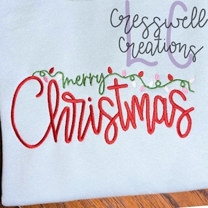 May include: A light blue sweatshirt with the embroidered words "Merry Christmas" in red script. The word "merry" is in green script, and the design includes a string of Christmas lights.