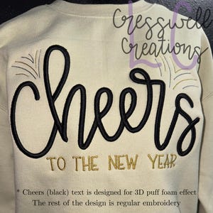 May include: A cream-colored sweatshirt with the word "Cheers" in large, black, puffy letters. Below, the phrase "TO THE NEW YEAR" is embroidered in gold. The design includes gold and black accents, celebrating the new year.