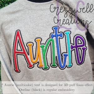 May include: A gray sweatshirt with the word "Auntie" embroidered in rainbow colors. The text is outlined in black. The text is designed for a 3D puff foam effect.  "Auntie (multicolor) text is designed for 3D puff foam effect. Outline (black) is regular embroidery."