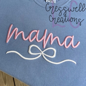 May include: A light blue t-shirt with the word "mama" embroidered in pink and a white bow underneath. The text "Cresswell Creations" is also embroidered on the shirt.
