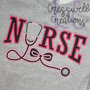 Puede incluir: Una sudadera gris con la palabra "NURSE" en letras negras bordeadas de rosa. Un estetoscopio rosa y negro con un lazo está bordado debajo de la palabra.