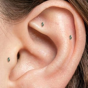 Op de afbeelding: Close-up van een oor met drie kleine, ovaalvormige, groene edelsteen oorbellen. De oorbellen zijn gezet in een zilver of wit metaal. Het oor is licht getint en de achtergrond is wazig.