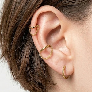May include: Three gold-toned ear accessories consisting of two ear cuffs with attached delicate chains and one small hoop earring. The upper ear cuff features a thin band with a chain, the middle cuff is slightly thicker with a chain, and the bottom is a plain hoop.