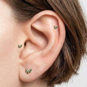May include: Close-up of three gold earrings with a leaf design. Each earring features small green gemstones. The earrings are displayed on an ear, showcasing the jewelry's placement and style.