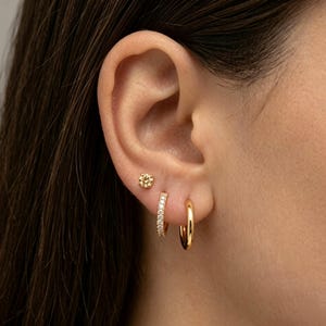May include: A set of three gold-toned earrings consisting of one small stud earring with a floral-shaped center, one small hoop earring encrusted with clear stones, and one plain, smooth gold-toned hoop earring worn in the earlobe.