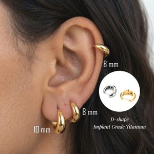 May include: D-shape hoop earrings made of implant grade titanium. The earrings are shown in gold and silver tones. The image displays sizes of 8 mm and 10 mm. The jewellery features a smooth, polished finish and a hinged closure mechanism for secure wear.