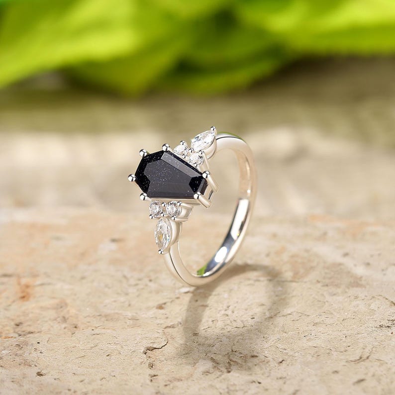 May include: A silver ring with a black coffin-shaped gemstone and smaller clear gemstones. The ring is set on a white background.