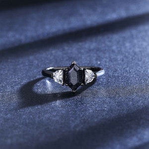 May include: A black ring with a large, dark, octagonal-shaped gemstone surrounded by two smaller, clear, triangular-shaped gemstones.