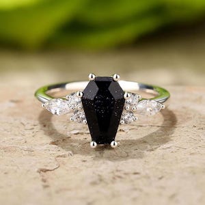 May include: A silver ring with a black coffin-shaped gemstone and smaller clear gemstones on the band.