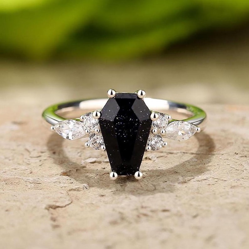 May include: A silver ring with a black coffin-shaped gemstone and smaller clear gemstones on the band.