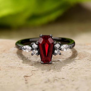 Könnte beinhalten: Schwarzer Ring mit einem roten, sargförmigen Edelstein und kleinen weißen Edelsteinen auf der Ringschiene.