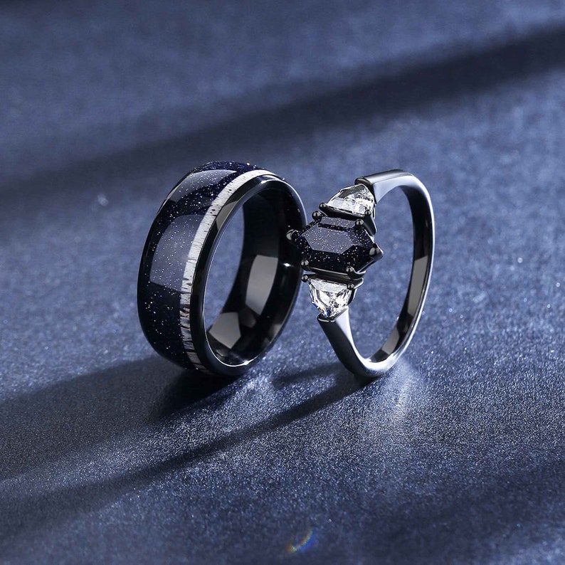 Op de afbeelding: Een paar zwarte trouwringen. De ring aan de linkerkant heeft een blauwe en witte gespikkelde afwerking. De ring aan de rechterkant heeft een zwarte edelsteen en twee kleinere witte edelstenen.