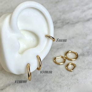 Op de afbeelding: Een set goudkleurige oorringen gepresenteerd op een wit oormodel. De ringen zijn gelabeld met de maten 8mm, 10mm en 12mm. Drie ringen zitten in het oormodel, terwijl twee extra losse ringen naast het model zijn geplaatst.