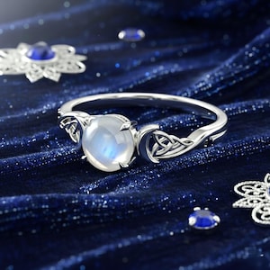 May include: A silver ring with a large, oval, iridescent moonstone. The ring features Celtic knotwork and crescent moon designs on the band. Small blue gemstones are set into the band. The ring is displayed on a dark blue velvet surface.