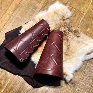 May include: Two brown leather arm guards with a diamond pattern and laces. The arm guards are on a brown fur surface.