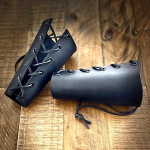 Two handcrafted black leather bracers featuring black eyelets and lacing, displayed on a rustic wooden surface. Ideal for medieval or Viking costumes, LARP, cosplay, or Renaissance fairs. Made from durable leather for comfort and authenticity.