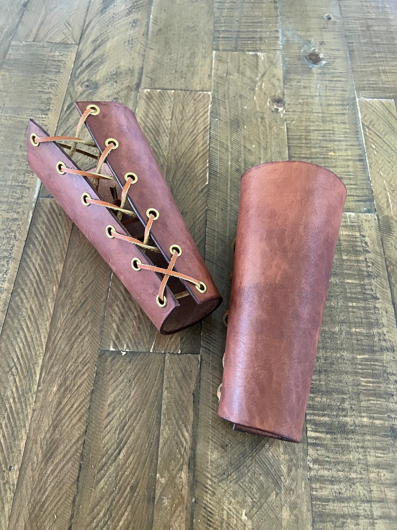Pair of handcrafted leather bracers with a rich brown finish and brass eyelets, laced with leather cord, displayed on a rustic wooden surface. Ideal for medieval costumes, LARP, Renaissance fairs, or cosplay.