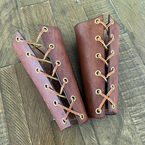 Pair of handcrafted leather bracers with a rich brown finish and brass eyelets, laced with leather cord, displayed on a rustic wooden surface. Ideal for medieval costumes, LARP, Renaissance fairs, or cosplay.