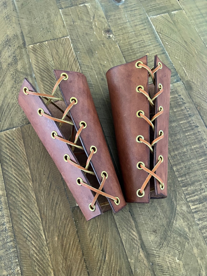 Pair of handcrafted leather bracers with a rich brown finish and brass eyelets, laced with leather cord, displayed on a rustic wooden surface. Ideal for medieval costumes, LARP, Renaissance fairs, or cosplay.