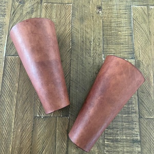 Pair of handcrafted leather bracers with a rich brown finish and brass eyelets, laced with leather cord, displayed on a rustic wooden surface. Ideal for medieval costumes, LARP, Renaissance fairs, or cosplay.
