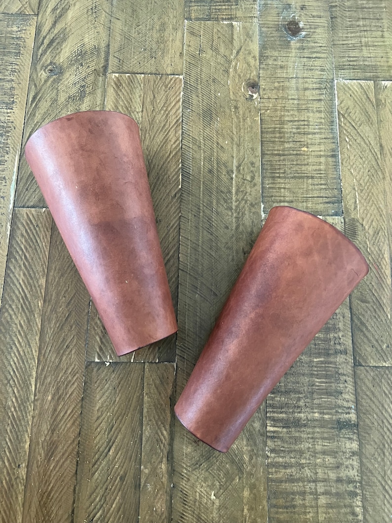 Pair of handcrafted leather bracers with a rich brown finish and brass eyelets, laced with leather cord, displayed on a rustic wooden surface. Ideal for medieval costumes, LARP, Renaissance fairs, or cosplay.