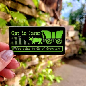 May include: Black sticker with green pixelated text and graphics. The text reads "Get in loser" above a covered wagon and animal graphic. Below, the text says "We're going to die of dysentery."