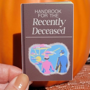 May include: A small, brown book with the title "Handbook for the Recently Deceased" in white lettering. The cover features a colorful illustration of two figures in front of a sunset. The book is held in a person's hand.