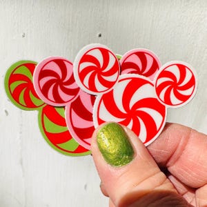 Op de afbeelding: Een verzameling kleurrijke, ronde stickers met kerstthema. De stickers hebben rood-witte spiraalontwerpen, sommige in roze en groen. De stickers worden vastgehouden door een hand met een groen geverfde vingernagel.