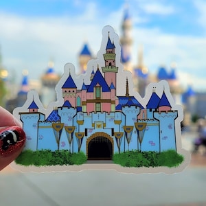 May include: A clear sticker of a pink and blue castle with gold accents. The castle has a large, arched entrance and is surrounded by green bushes.
