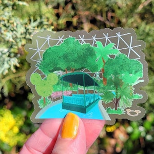 May include: A sticker depicting a green boat with a canopy, surrounded by green trees and a white lattice structure. The sticker is on a hand.