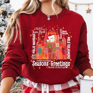 Peut inclure: Sweat-shirt rouge de Noël avec une illustration colorée d'un château et le texte "Seasons Greetings" dans une police festive. Le château est décoré de lumières et d'ornements, et le texte est entouré d'étoiles et de flocons de neige. Le sweat-shirt présente également des salutations dans plusieurs langues, notamment "Joyeux Noël", "Feliz Navidad", "Buon Natale", "Namaste" et "Frohe Feiertage".