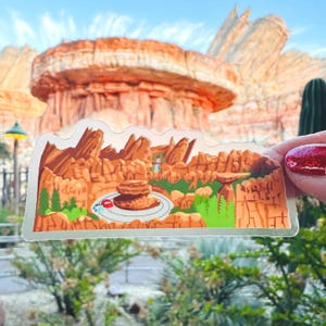 May include: A die-cut vinyl sticker featuring a colorful illustration of a desert landscape with orange rock formations, green pine trees, and a small red car parked on a circular gray road, set against a backdrop of a large red rock mesa.