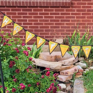 Może przedstawiać: Żółty baner z napisem "HAPPY" w białych literach, zawieszony na czarnym sznurku. Baner ma zieloną obwódkę i jest umieszczony na tle ceglanej ściany, kwiatów i kamiennych schodów.