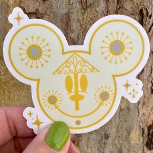May include: A die-cut sticker in the shape of Mickey Mouse's head, featuring a yellow and white design. The sticker has a stylized face with sunburst details and a decorative house shape. The sticker is held against a wood background.