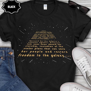 Puede incluir: Camiseta negra con un diseño de texto dorado que presenta una escena espacial y texto de una película de ciencia ficción. El texto dice: "Perseguida por los siniestros agentes del Imperio, la princesa Leia..."