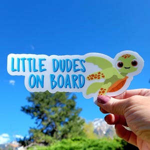 May include: A die-cut sticker featuring a cartoon sea turtle with a light green body and orange shell accents. The text "LITTLE DUDES ON BOARD" is printed in a bold, blue, sans-serif font to the left of the turtle illustration on a white background.