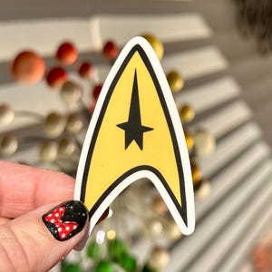 May include: A yellow and black Star Trek badge sticker. The sticker has a yellow background with a black star and arrow design. The sticker is held by a hand with a nail design.
