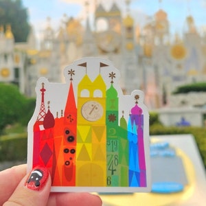 Könnte beinhalten: Ein bunter Aufkleber mit einem Schloss in Regenbogenfarben. Das Schloss hat ein Zifferblatt mit der Zahl 4. Der Aufkleber befindet sich vor einem verschwommenen Hintergrund eines Freizeitparks.