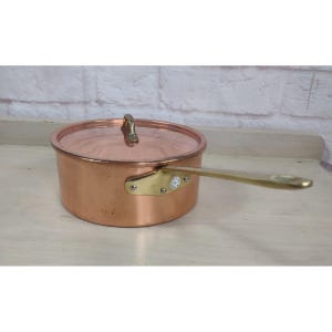 May include: A round copper saucepan with a matching lid and a long brass handle. The handle is attached to the side of the pan with a brass plate secured by three rivets. The lid features a small brass handle centered on the top surface.