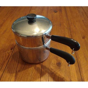 May include: A stainless steel double boiler with a lid and two black handles. The pot is sitting on a wooden surface. The lid has a black knob.