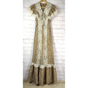 May include: A vintage brown floral print maxi dress with white lace trim. The dress features short sleeves, a V-neckline with a tie, and a tiered skirt. The dress is hanging on a wooden hanger against a white brick wall.