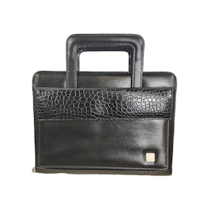 May include: A black structured briefcase with a rectangular top handle. The front features a smooth leather-like finish with a horizontal panel of embossed crocodile-patterned texture and a small square gold-toned metal emblem in the corner.