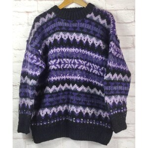 May include: A cozy, oversized sweater in shades of purple, black, and white. The sweater features a crew neck and ribbed cuffs, with a geometric pattern across the chest and sleeves. The sweater is displayed on a wooden hanger.