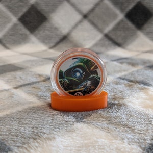 May include: A small, round, orange plastic container with a clear lid. The lid has a printed image of a green and blue character with a white eye.